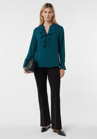 Teal blouse with ruffled neckline, long sleeves, and a smooth texture; paired with black flared pants and a small black handbag.
