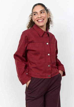 Vrouw glimlachend, draagt een donkerrode met knopen gesloten jas en donkere broek, handen in de zakken, kijkt opzij tegen een effen witte achtergrond.