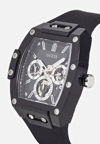 Montre en caoutchouc noir avec un boîtier carré, des accents argentés et un cadran transparent présentant plusieurs sous-cadrans et des marqueurs blancs.