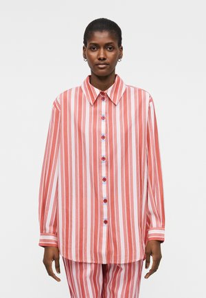 Žena u širokoj crveno-bijeloj vertikalno prugastoj košulji s okovratnikom i gumbima, stoji pred jednostavnom svijetlom pozadinom.