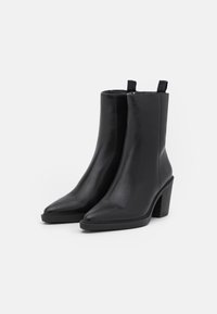 Botas de piel negra hasta el tobillo con punta afilada y un tacón alto y grueso. Presentan tiradores para facilitar su uso. Textura suave y pulida en todo el zapato.