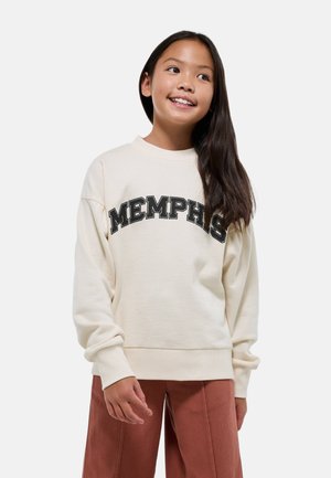 Jeune fille aux longs cheveux bruns portant un sweat-shirt crème avec le texte "MEMPHIS" et un pantalon couleur rouille, souriante et regardant sur le côté.