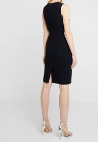 Svart ärmlös klänning med en strukturerad passform, försedd med dragkedja i ryggen och en liten slits bak, tillsammans med silverfärgade klacksandaler.