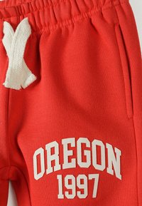 Červené teplákové nohavice s elastickým pásom, bielou šnúrkou, bočnými vreckami a potlačou "OREGON 1997" v tučných bielych písmenách. Hladký povrch.