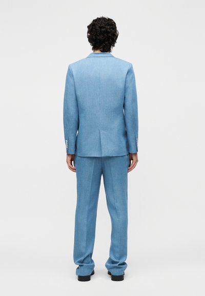 Homme aux cheveux bouclés portant un costume bleu clair sur mesure et des chaussures noires, debout de dos sur un fond blanc uni.
