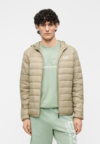 Geacă matlasată bej cu glugă, cu un design matlasat orizontal, asortată cu un hanorac verde deschis și pantaloni de trening pe măsură.