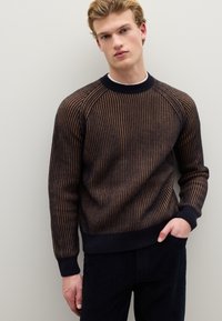 Gestrickter Pullover in Dunkelblau mit senkrechten braunen Streifen. Verfügt über einen gerippten Kragen und Bündchen. Modell trägt ihn mit Händen in den Taschen.