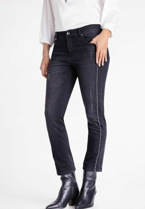 Woman wearing black skinny jeans with silver studs along outer seams and black ankle boots, standing with hand in pocket.