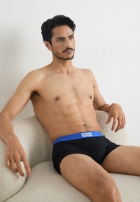 Black boxer shorts with a blue waistband labeled "HUGO," worn by a seated individual against a light background. Smooth texture, fitted design.