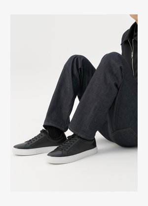 Ténis de couro preto com solas de borracha brancas, apresentando atacadores redondos e um design minimalista, combinados com jeans de ganga escuros.