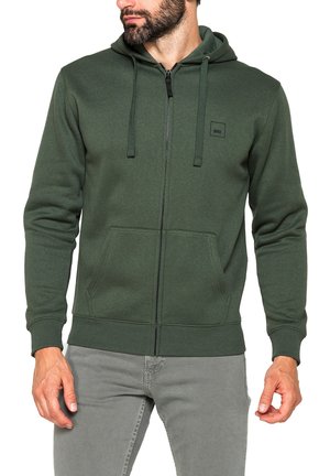 Uomo che indossa una felpa con cappuccio verde scuro con zip e tasche anteriori, pantaloni grigio chiaro, in piedi su sfondo bianco.
