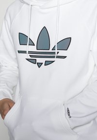 Vit hoodie med en teal och svart adidas-logotyp, känguruficka och snörhuva. Mjuk tyg med ribbade muddar och nederkant.