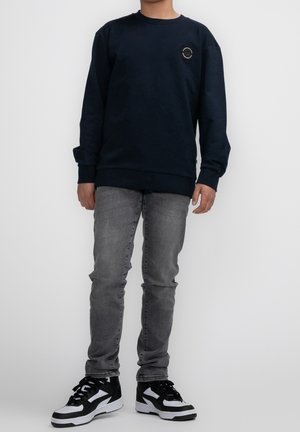 Felpa navy con colletto tondo e una piccola patch con logo. Indossata con jeans grigi e sneakers bianco e nere. Design semplice.