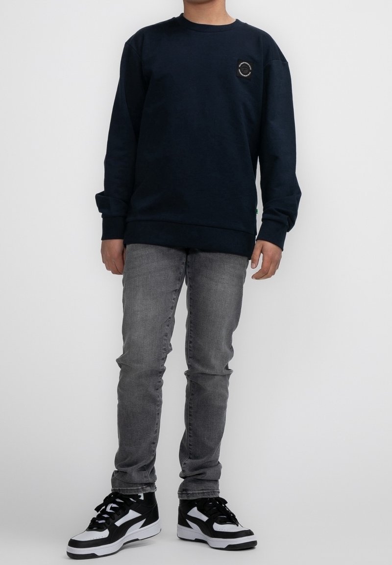 Sweat-shirt bleu marine avec un col rond et un petit patch logo. Porté avec un jean gris et des baskets noir et blanc. Design simple.