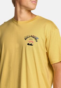 T-shirt di cotone gialla con un design grafico frontale che presenta testo e motivi a onda. Maniche corte e collo tondo classico.