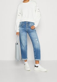 Bílá mikina s dlouhými rukávy a vykrojenými detaily, světle modré obnošené džíny s vysokým pasem, bílé tenisky s černými akcenty a šedá taška.