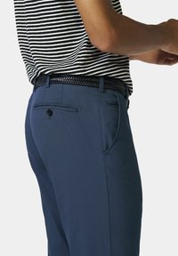 Navy blauwe broek met een gladde textuur, met een geweven zwarte riem. Achterzak met een knoopdetail. Gestreept shirt dat gedeeltelijk zichtbaar is.