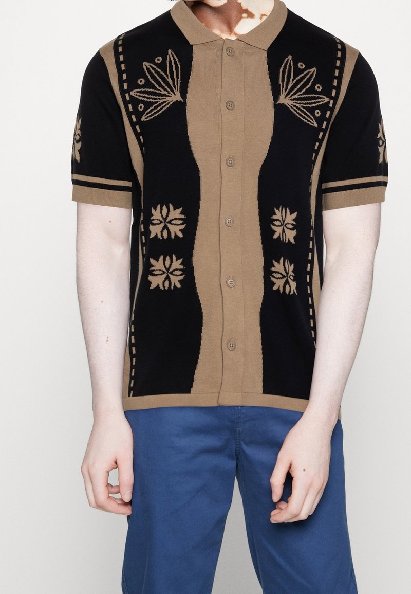 Personne portant une chemise à manches courtes boutonnée avec des motifs floraux noirs et beiges, associée à un pantalon bleu, debout devant un fond uni.