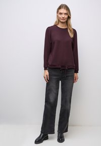 Bordo pitkähihainen collegepaita pyöreällä kaula-aukolla, kerättyjen olkien ja säädettävän nyörin reunassa, yhdistettynä mustiin leveisiin farkkuihin ja nilkkasaappaisiin.