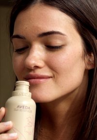 Bouteille de produit capillaire Aveda, beige avec logo en relief et motif floral, tenue près du visage d'une personne à la peau lisse et aux cheveux longs.