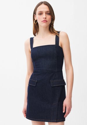 Donna che indossa un vestito di jeans smanicato blu scuro con scollo quadrato e tasche con patta, su sfondo chiaro e semplice.