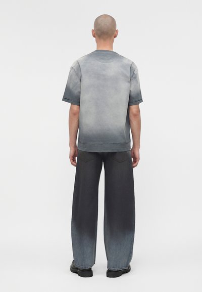 Άνδρας με ξυρισμένο κεφάλι που φοράει ξεβαμμένο γκρι μπλουζάκι με κοντά μανίκια και φαρδιά τζιν με διαβάθμιση από μαύρο σε γκρι, στέκεται μπροστά σε ένα μονόχρωμο φόντο.