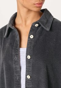 Moteris vilki tamsiai pilką kordurovinę marškinių su baltomis sagomis virš šviesiai pilko viršutinio drabužio, matoma kaklo ir dalies apatinių veido bruožų.