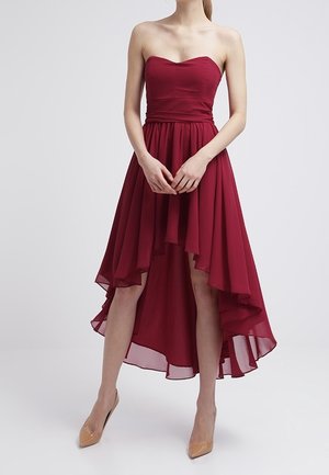 Femme portant une robe bordeaux sans manches ni bretelles, à jupe fluide asymétrique, et des talons pointus couleur nude, debout devant un fond uni.