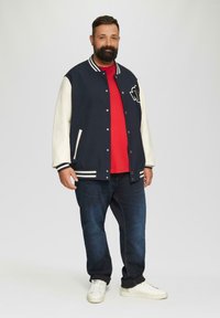 Marineblauwe en roomkleurige varsity-jack met witte strepen, een rode T-shirt, donkere jeans en witte sneakers. Het jack is voorzien van een logo en zakken.