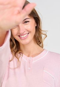 Mujer sonriente con cabello ondulado vestida con una blusa rosa, levantando su mano hacia la cámara y cubriendo parcialmente su rostro.