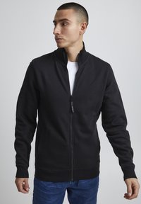 Veste zippée noire en coton lisse avec col montant, manches longues et poches latérales. Associée à un t-shirt blanc en dessous.