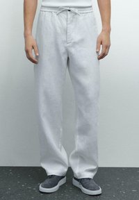 Pantalons en lin blanc à coupe décontractée, avec une taille élastique et un cordon de serrage. Portés avec des chaussures en suède grises sur une surface texturée sombre.