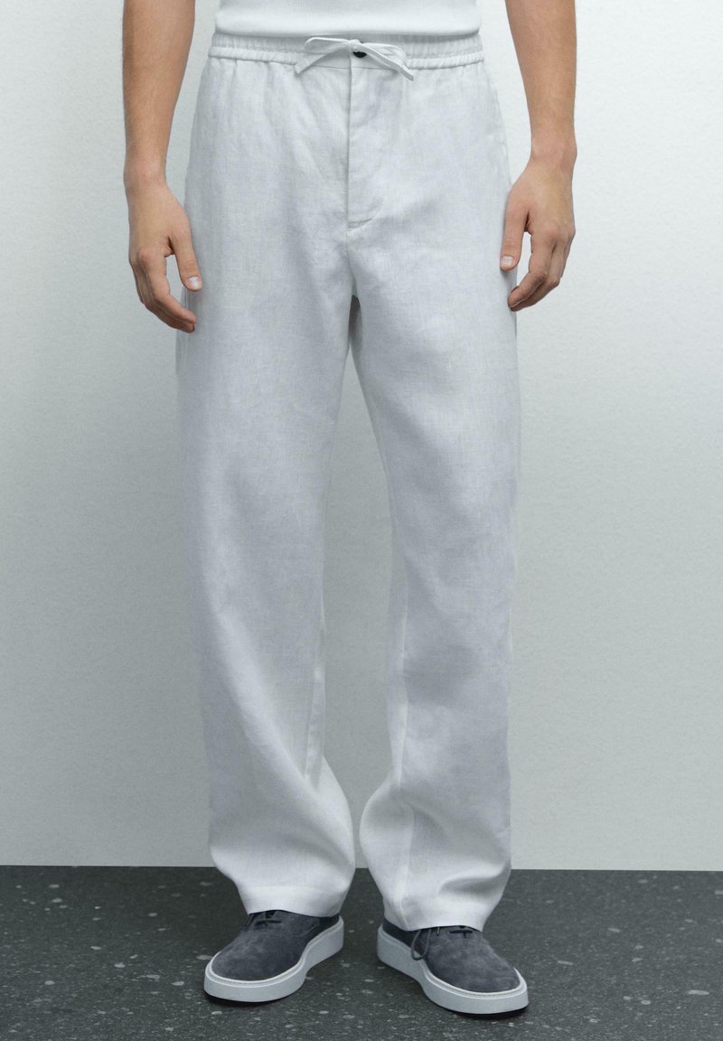Pantalons en lin blanc à coupe décontractée, avec une taille élastique et un cordon de serrage. Portés avec des chaussures en suède grises sur une surface texturée sombre.