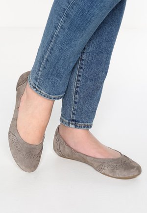 Pieds en jean bleu portant des ballerines en daim taupe avec des perforations décoratives sur un sol blanc.