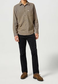 Geruite bruine overhemd met lange mouwen, een zak en knoopsluiting, gecombineerd met zwarte broek en bruine suède schoenen.