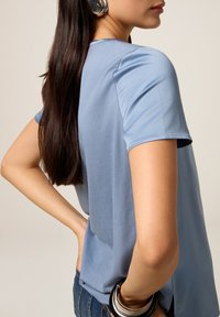 Tricou albastru, cu mâneci scurte, dintr-un material ușor, cu un guler rotund și o croială lejeră, asortat cu blugi și o brățară.