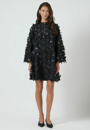 Mujer con vestido negro hasta la rodilla con apliques florales 3D y lentejuelas, combinado con zapatos de tacón negros de punta afilada, de pie sobre un fondo liso.