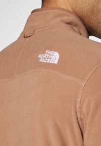 Barna fleece dzseki magas gallérral, fehér hímzett logóval a hátsó felső részen, és látható varrási részletekkel. Puha tapintású.