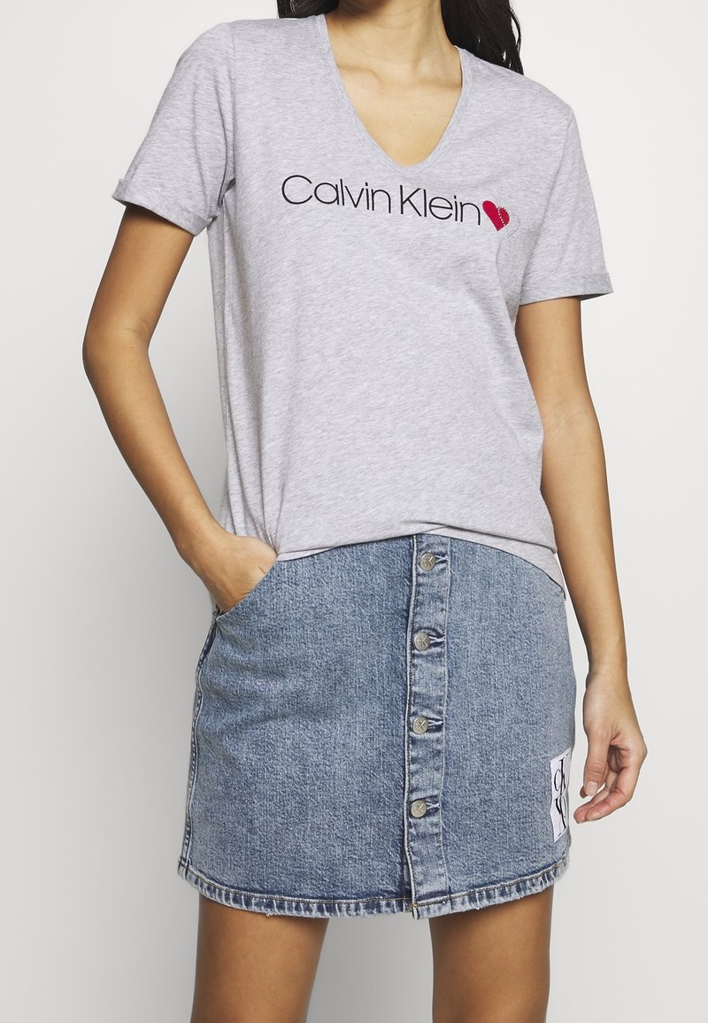 Šedé tričko s výstřihem do V a černým logem "Calvin Klein", spárované s modrou džínovou sukní s předními knoflíky a bočními kapsami.