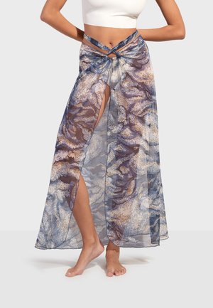 Kobieta ubrana w przezroczystą, wzorzystą spódnicę plażową z przodem związanym w węzeł i bocznym rozcięciem, w połączeniu z białym crop topem. Widoczne bose stopy.