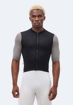 Szőke hajú férfi, akinek fekete, testre simuló kerékpáros mez van rövid mintás ujjakkal és fehér párnázott kerékpáros nadrág, előre nézve áll.