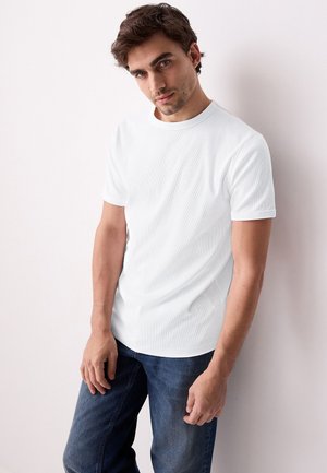 Junger Mann mit braunen Haaren, der ein schlichtes weißes Kurzarm-T-Shirt und blaue Jeans trägt und sich an eine weiße Wand lehnt.
