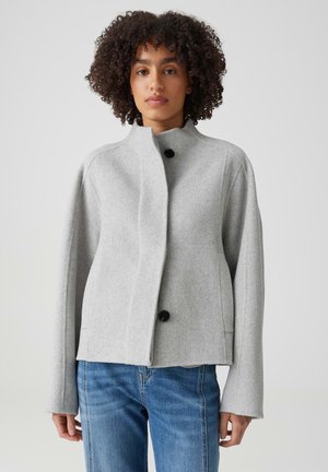 Woman with curly hair wearing a light gray buttoned jacket and blue jeans, standing against a plain light background.