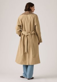 Γυναίκα που φοράει μπεζ παλτό με ζώνη, μπλε τζιν και μαύρα παπούτσια, στέκεται γυρισμένη πλάτη σε απλό φόντο.