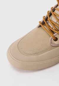 Zbliżenie na beżowy czubek trampka z teksturowaną gumową podeszwą i sznurówkami w pomarańczowym akcentem na jasnoszarym tle.