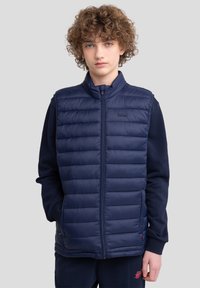 Gilet rembourré bleu marine avec un col haut, un matelassage horizontal et une fermeture éclair à l'avant. Porté par-dessus un sweat-shirt bleu marine, avec des poches latérales et un détail de logo.