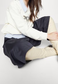 Vit sweatshirt med en fågeldesign, lagd över en ljusblå randig skjorta, tillsammans med en svart kjol och beige leggings.