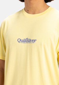 Camiseta de algodón color amarillo claro con un logo azul de Quiksilver y texto en el pecho. Manga corta y cuello redondo clásico.