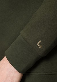 Rękaw bluzy w kolorze oliwkowym z teksturowanym wzorem dzianiny i małym, beżowym haftowanym logo na rękawie. Miękki, elastyczny materiał.