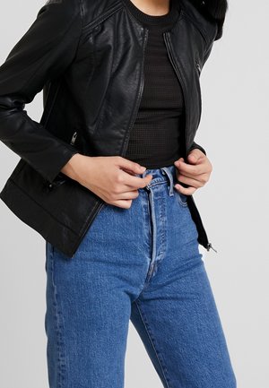 Personne portant une veste en cuir noire sur un haut texturé noir et un jean bleu taille haute, ajustant la fermeture éclair de la veste.